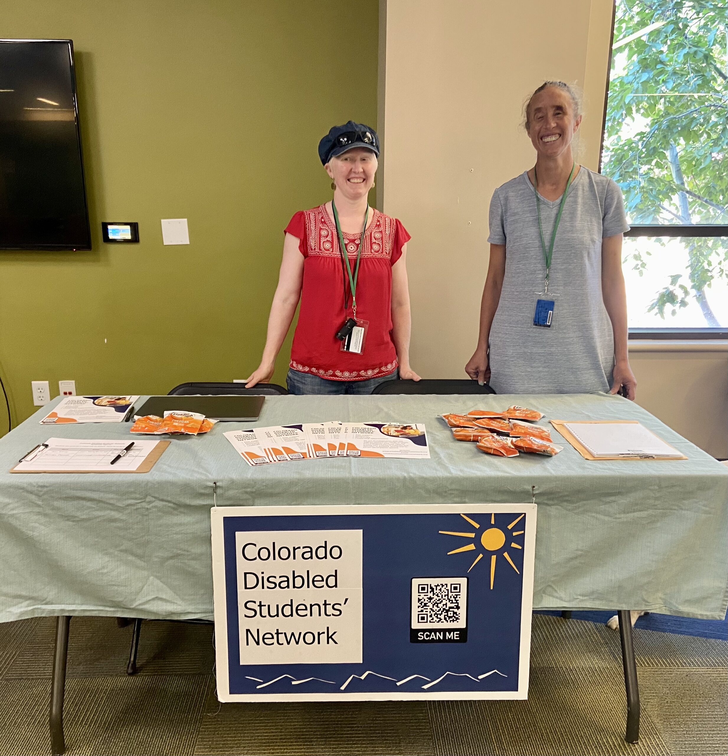 Anna and Amelia standing behind a table with flyers, snacks, and a poster board sign that says Colorado Disabled Students' Network.